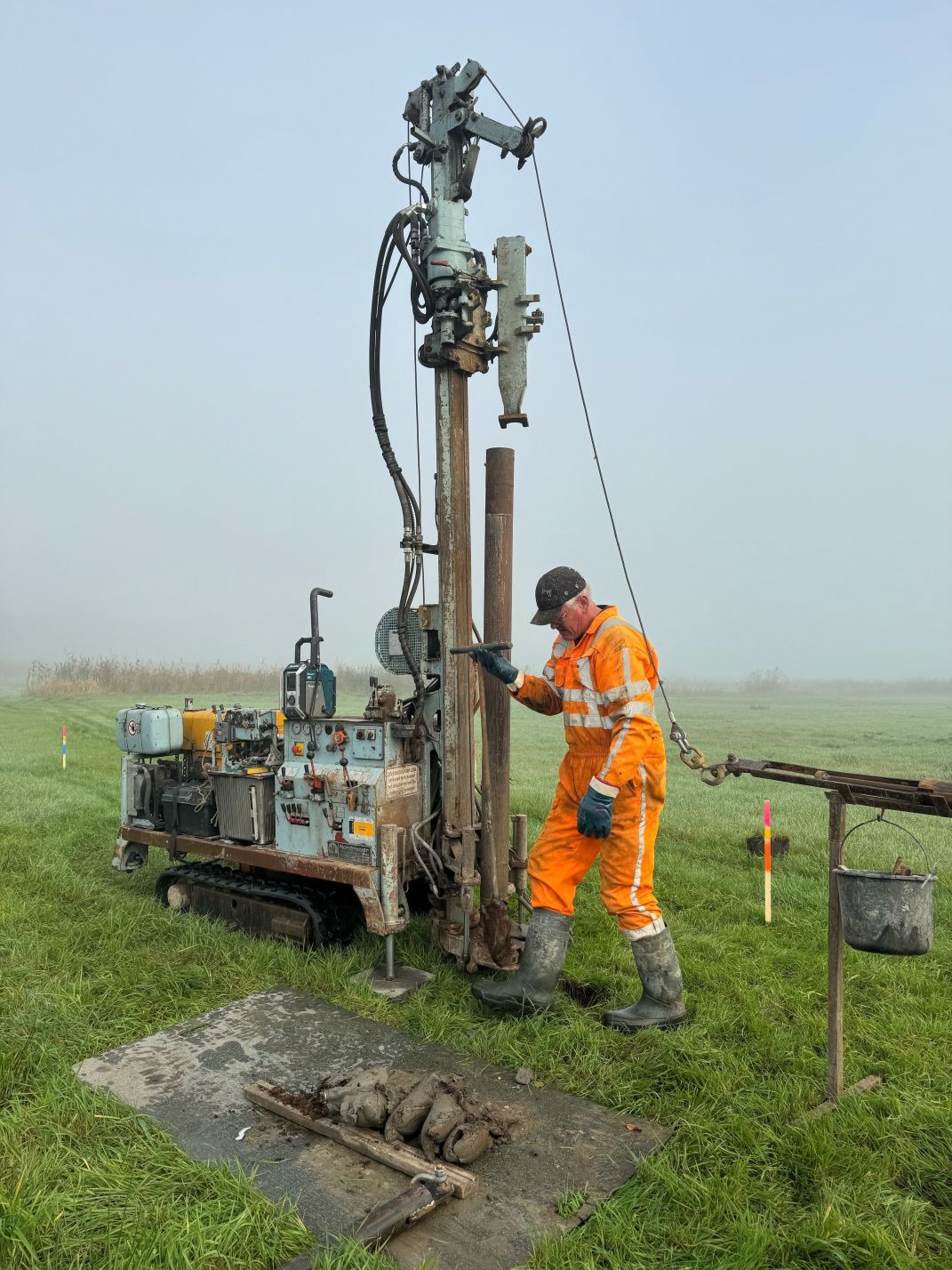 Onderzoeker bij voertuig op rupsbanden. Aan het voertuig is een boormachine bevestigd. Op de voorgrond liggen monsters van veengrond die uit de bodem zijn gehaald. 
