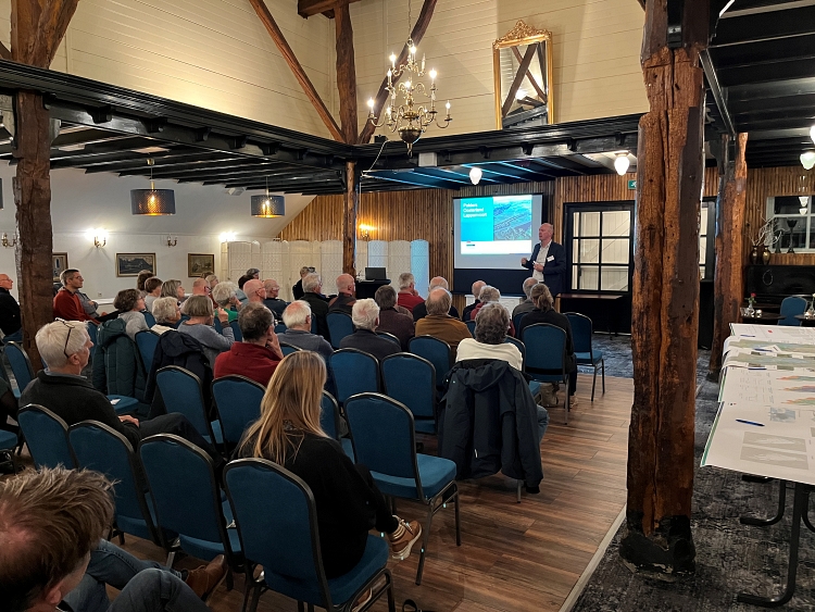 Een presentator voor een projectiescherm, met een zaal vol luisterende bezoekers.