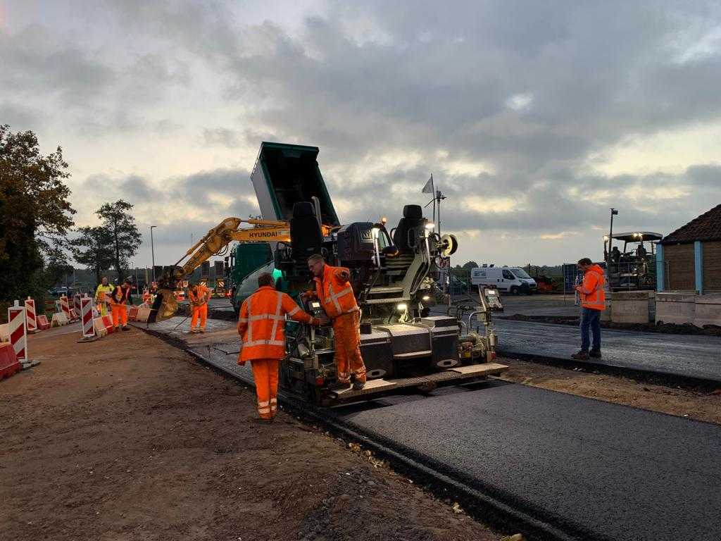 Met schemer zijn meerdere mensen in oranje werkkleding bezig met asfalteren.
