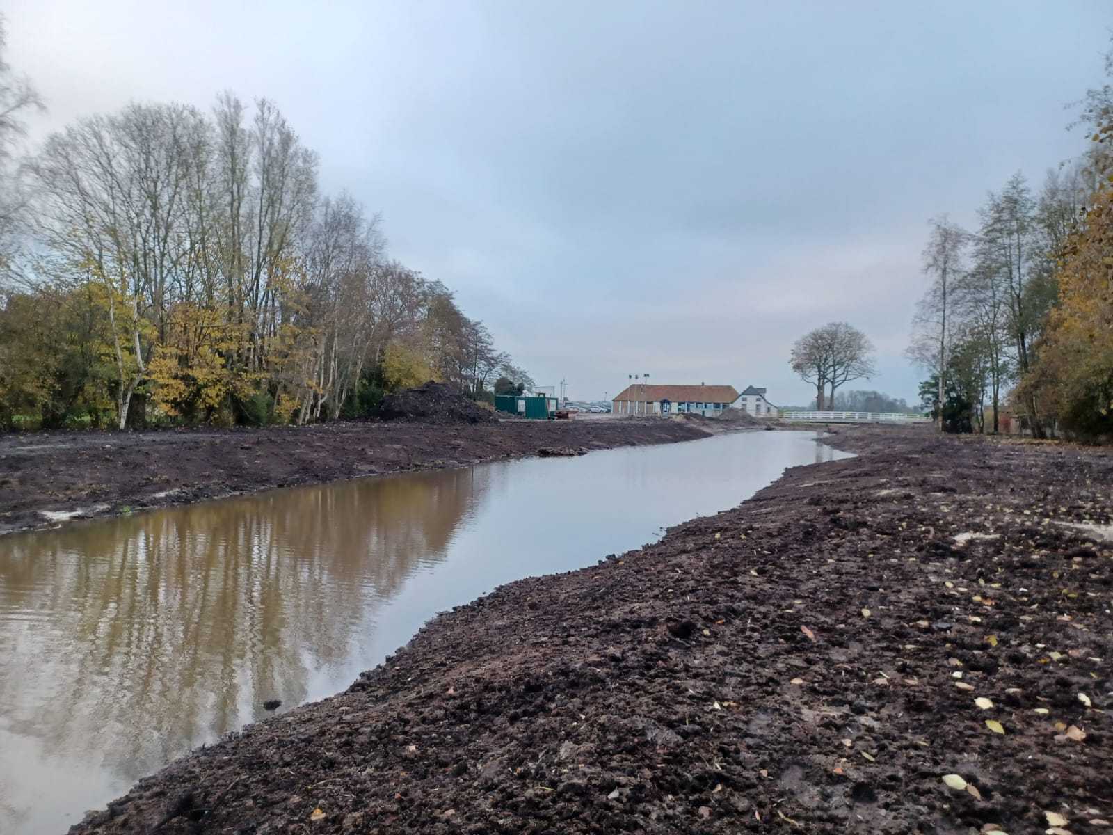 Slenk tussen twee kades, met bomen en een gebouw in de achtergrond.